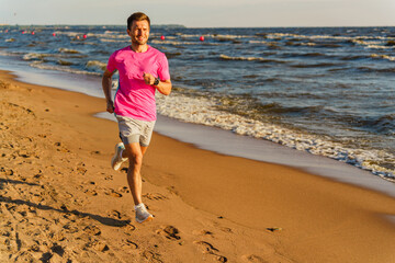 A man runs along a sunny beach near the waves, wearing a pink shirt and white shoes, smiling as he...