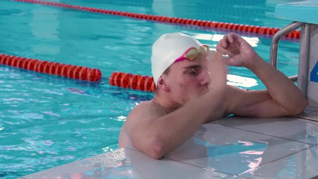 Tired, but happy with the swim and training, a young male swimmer athlete in a swimming pool. The guy emerges from the water and shows an approving gesture. The concept of sports and health care.