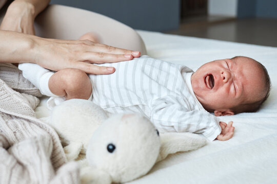 Unrecognizable woman massaging tummy of her crying sick son trying to relieve colic infant baby wearing striped bodysuit lying on bed with fluffy toy