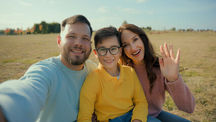 Happy European smiling family blogger man woman parents son little boy child kid mom dad mobile phone looking camera streaming recording video call greeting waving hands trip outside rural meadow park