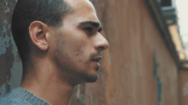 20-year-old man with short hair and gray sweater, standing against a wall in a suburban courtyard, smoking a cigarette with a serious look on his face.