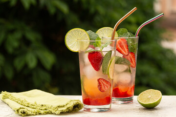 Refreshing summer cocktail with strawberries, lime, mint leaves, ice cubes, strawberry mojito. Cold fresh drink  in a glass, green outdoor  on background.
