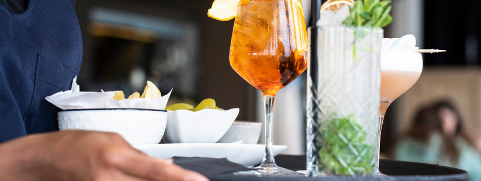 Waiter serving refreshing cocktails and snacks on a tray in a stylish bar. The drinks include an orange spritz and a mint garnished cocktail, perfect for a relaxing indoor setting.