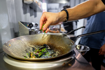 Close-up of a person’s hand sprinkling herbs into a wok with food, capturing the essence of cooking