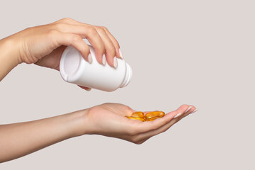 A woman pours vitamins or capsules into her hand from a white plastic bottle, isolated on a beige background. The girl takes omega for beautiful skin and nails. Beauty and health care concept