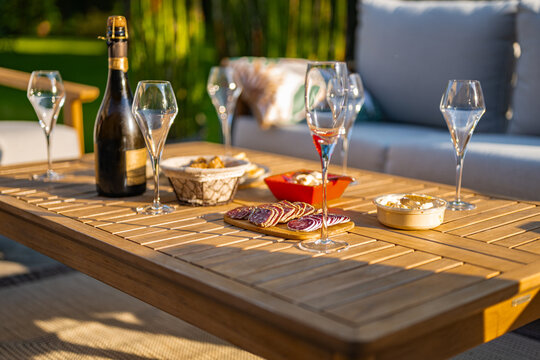 Ap&eacute;ritif servi sur table de jardin en &eacute;t&eacute;, au coucher du soleil, avec une bouteille de champagne et ses fl&ucirc;tes, du saucisson, des biscuits, du pain grill&eacute;, du houmous et du tarama.