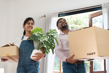 Indian Couple Moving into New Home, Carrying Boxes and Plant, Smiling and Excited, Bright and Modern Interior, Relocation and New Beginnings, Happy Homeowners, Diverse and Contemporary Lifestyle