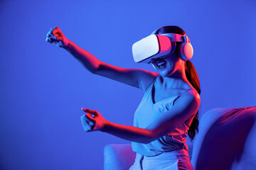 Smart Female sitting on sofa surrounded by neon light wear VR headset connecting metaverse,...