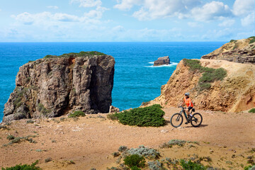 happy active senior woman cycling on the the rocky cliffs of the Vicentina coast of  Algarve,...