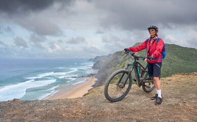 happy active senior woman cycling on the the rocky cliffs of the Vicentina coast of  Algarve,...