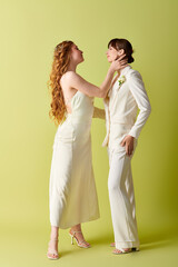 A lesbian couple dressed in white wedding attire stand facing each other against a green backdrop.