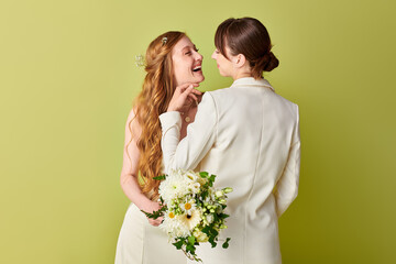 Two young women in white wedding attire smile at each other against a green backdrop.