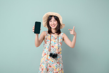 Travel promotion screen concept. Happy Asian woman wearing casual dress and hat with camera showing mobile phone screen isolated on pastel green background.