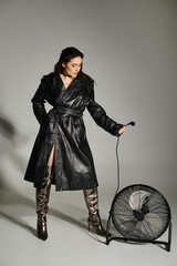 Plus size woman exudes sophistication in trench coat, showcasing fan in striking pose on gray backdrop.