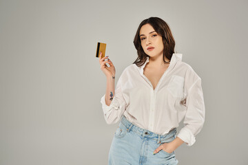 Beautiful plus-size woman in a white shirt confidently holding a gold card against a gray backdrop.