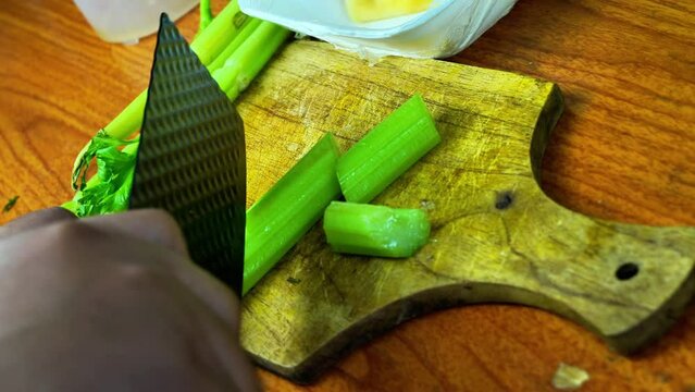 A person chops celery stalks and pineapple on a wooden cutting board.