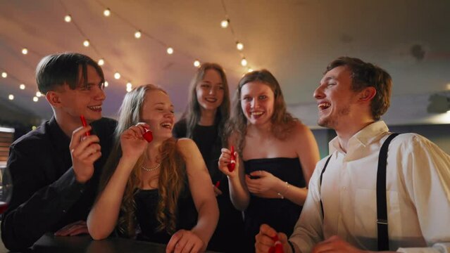 People celebrating b-day, blowing noisemakers whistles, congratulation party. Portrait of happy five best friends cheering and greeting, merry birthday party, gathering in festival in nightclub