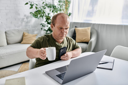 A man with inclusivity in casual attire sits at a table in his home, enjoying a cup of coffee while using a laptop and smartphone.