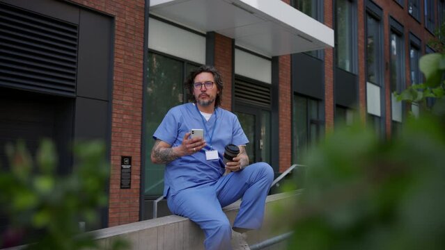 Healthcare worker taking a break, scrolling a phone and drinking coffee outdoors, in front of the hospital.
