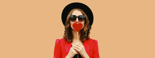 Beautiful stylish woman blowing kiss with sweet red heart shaped lollipop on stick in black hat