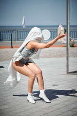 Young sexy girl in space silver micro skirt dancing with white silk scarf waving gracefully, female outdoor dance performance on seaside promenade creating an arousing outdoor spectacle