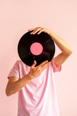 Portrait of man in pink shirt holding vinyl record near his face on pink background.