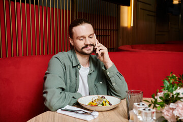 A man, seated at a table, engages in conversation on a cell phone in a modern cafe setting.