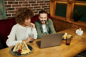 An African American woman and a man sit at a table in a modern cafe, busily focused on the laptop...