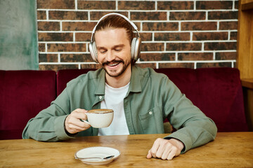 A man of African descent sits at a cafe table, sipping coffee and listening to music through...