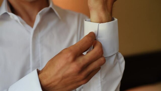 Smiling Caucasian man looking at mirror, combing hair with fingers, getting ready for new wedding day. Stylish young man in blue suit adjusts his cufflinks buttons white shirt putting on jacket. 