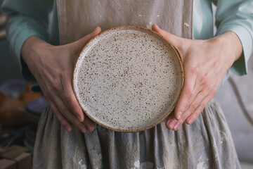 Ceramist woman in her workshop holds a handmade ceramic plate in her hands in a work apron.