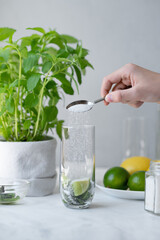 Hand holding spoon pouring sugar in glass with lime and mint leaves, process of mojito cocktail drink preparing, making homemade refreshing summer drink