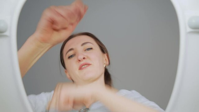 Cheerful woman dances and rejoices, peering into open toilet lid, giving thumbs-up, indicating successful completion of restroom cleaning. View from toilet bottom. Cleanliness and hygiene concept