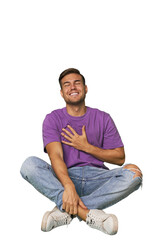 Casual man sitting on floor laughs out loudly keeping hand on chest.