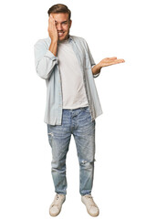 Full body portrait of a man in studio holds copy space on a palm, keep hand over cheek. Amazed and delighted.