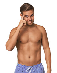 Naklejka premium Young man shirtless in swimwear, studio pointing temple with finger, thinking, focused on a task.
