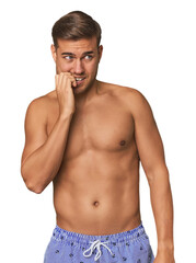 Young man shirtless in swimwear, studio biting fingernails, nervous and very anxious.