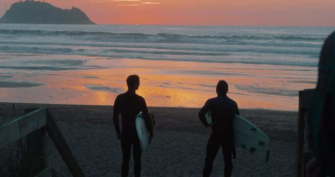 Silhouette of two surfers stand at beach at sunset overlooking waves, waiting for tide to change. After surf session or early morning warmup. Cinematic surfing destination