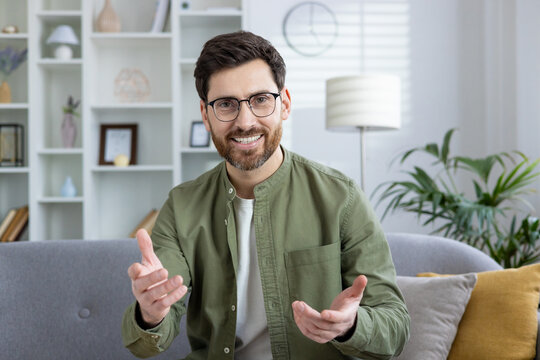 Smiling young man in shorts sitting on sofa at home and talking casually on video call, explaining while gesturing with hands to camera. - Powered by Adobe