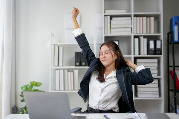 Young businesswoman in office stretching with eyes closed, appearing relaxed and relieved. Office environment with laptop, documents, and books.