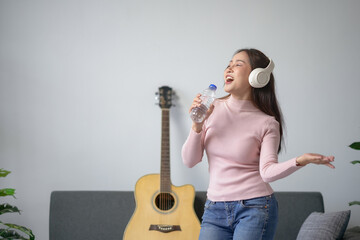 Young woman singing with headphones while holding a water bottle, guitar in background, having fun...