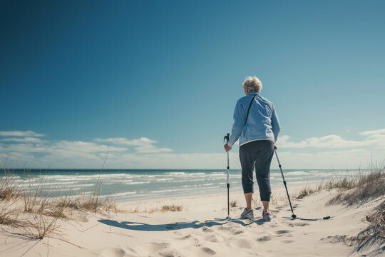 A woman practices Nordic walking on a sandy beach using trekking poles under the clear blue sky. Senior Citizens Day