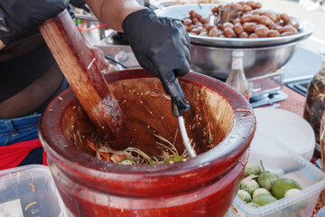 A close-up view of a person wearing black gloves using a wooden mortar and pestle to prepare traditional Thai papaya salad, also known as Som Tum, at a street food stall. 
