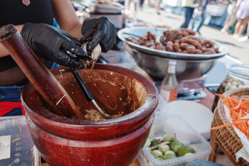 A close-up view of a person wearing black gloves using a wooden mortar and pestle to prepare traditional Thai papaya salad, also known as Som Tum, at a street food stall. 