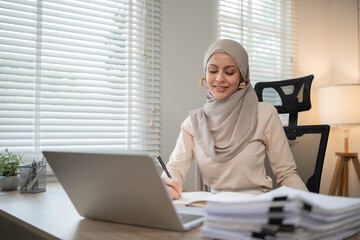 confident Muslim Woman Working at Home Office, Wearing Hijab, Using Laptop and Writing Notes, Modern Workspace with Natural Light, Professional and Productive Atmosphere