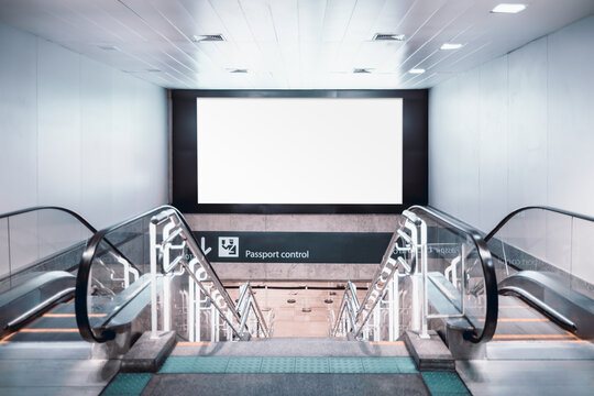 Two escalators and staircase leading down to a passport control area with a large blank billboard above for advertisement mockup. The scene is set in a modern, well-lit airport terminal interior - Powered by Adobe