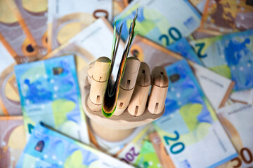 A wooden hand holds a stack of twenty Euro banknotes against a backdrop of additional twenty Euro notes