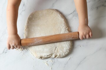 Little child rolling raw dough at white table, top view