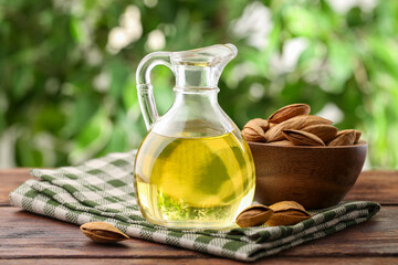 Almond oil in jug and nuts on wooden table against blurred green background, closeup