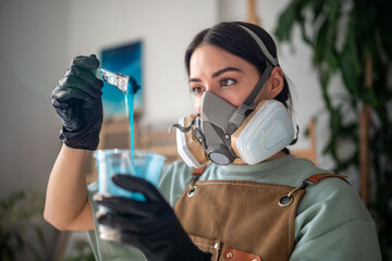 Attentive female artist wearing respirator and latex gloves tests blue epoxy resin composition for creative working in art studio. Focused female artist examines liquid material for seascape paintings - Powered by Adobe